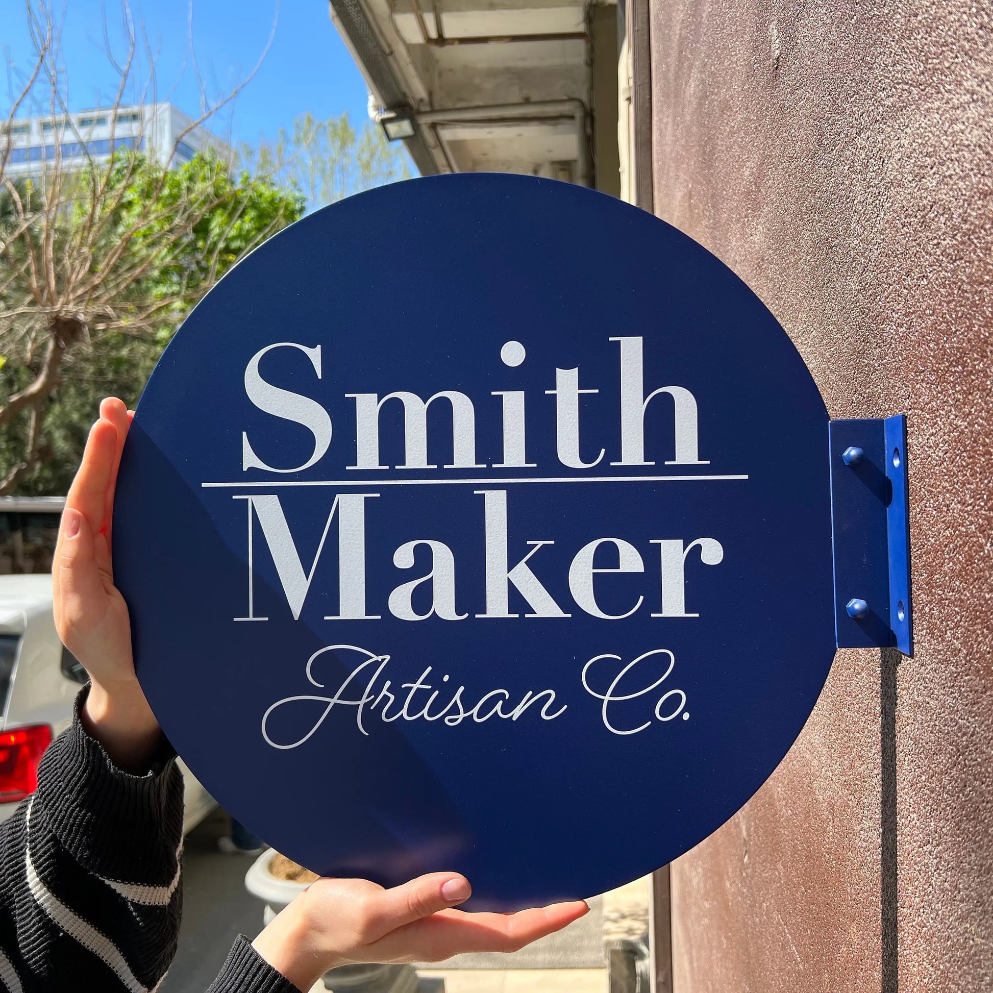 Round blue sign with 'Smith Maker Artisan Co.' text held by a person outdoors.